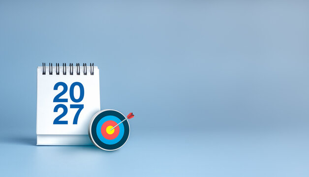 Desk calendar displaying 2027 beside 3d bullseye with arrow on blue banner background with copy space. Annual goals, deadline planning, strategic focus, business performance and achievement concept.