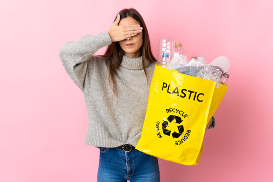 Young caucasian woman holding a bag full of plastic bottles to recycle isolated on pink background covering eyes by hands. Do not want to see something
