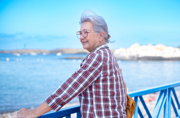 Fototapeta premium Senior adult woman enjoying a relaxing coastal vacation, smiling while looking at the calm blue sea, standing on a pier during retirement travel and leisure time
