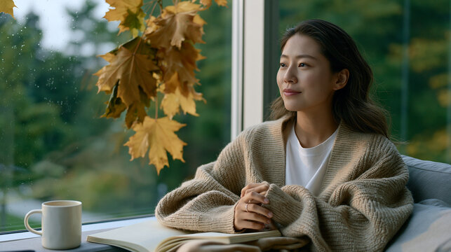 Graduate student wrapped in an oversized cardigan sitting by a rain-streaked window with a mug, open notebook, and fallen maple leaves pressed against the glass outside capturing