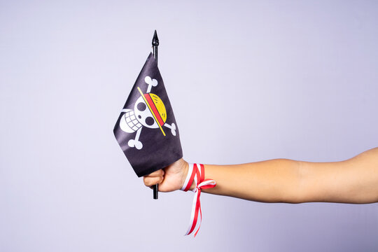Riau, August 1, 2026, Extended arm holding pirate flag "Jolly Roger" with skull and straw hat symbol, inspired by anime, Red and white ribbon tied around wrist. Isolated on plain background.