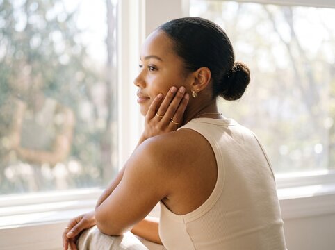 Serene young woman with hand on cheek, looking out a sunlit window in quiet contemplation, natural light illuminating her profile