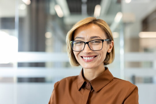 Professional woman wearing glasses, smiling brightly and looking at the camera, conveying success, confidence, and positive attitude in a modern corporate environment