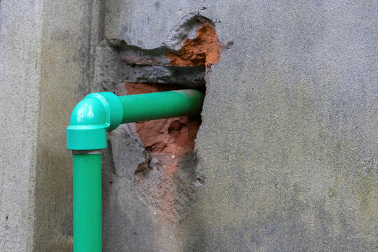 Green PVC plastic pipe passing through damaged hole in concrete building wall revealing red brick layer. Plumbing renovation and construction repair.