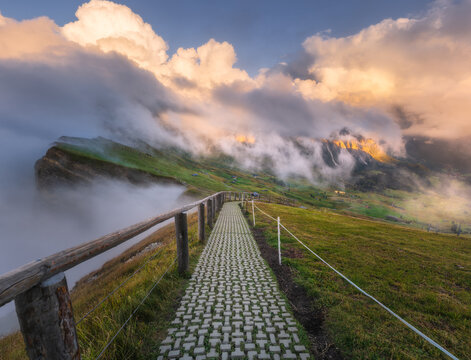 Paved path with wooden fence leading to mountain cliffs at sunset in Dolomites, Italy. High altitude alpine trail view with clouds and fog over green valley, sunset light on rocky mountain peak Seceda