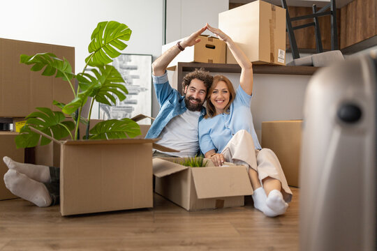 Couple sits together in a room full of moving boxes while making a heart shape with their hands