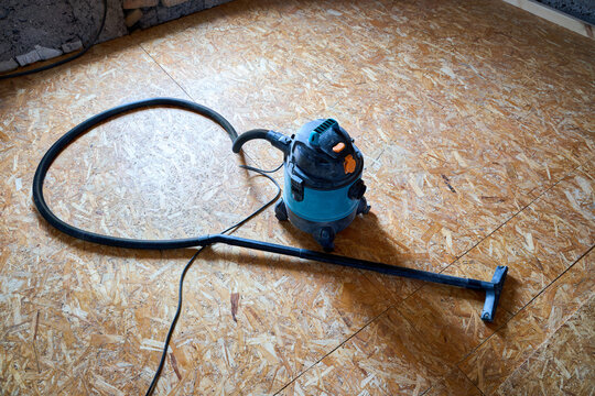 Industrial blue vacuum cleaner on wooden osb floor panels in room under renovation process. Dirty workshop power tool for professional dust cleaning service on construction site.