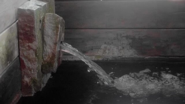 Hot spring thermal water pouring from stone spout into dark wooden spa bath with steam