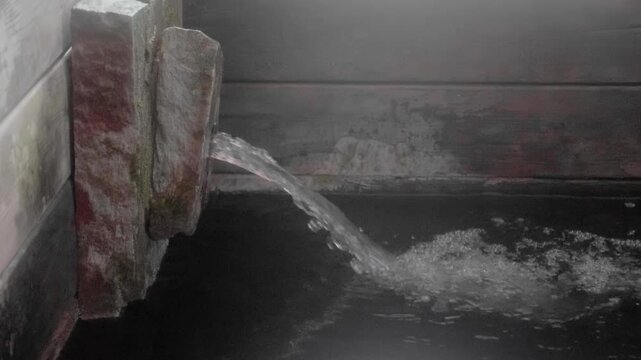 Hot spring water flowing from stone spout into wooden bath tub in traditional Japanese onsen spa resort