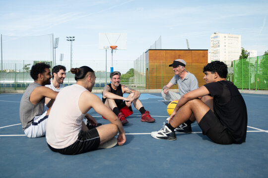 Group of diverse men sitting on a basketball court, laughing and talking in a huddle