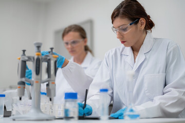 Fototapeta premium Professional female scientist writing research data on digital tablet in modern laboratory with colleague. Advanced medical biotechnology and commercial pharmaceutical testing equipment.