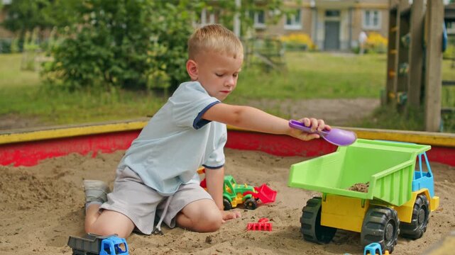 Small blonde child in a sandbox, digging in the sand with a toy shovel.  Kid having fun on a playground, child entertained in a sandpit with many toys. Little boy playing in a sandbox.