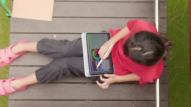 Top view of an Asian kid enjoying a relaxing hobby, drawing digital art on a tablet. Selective focus captures the elementary student spending casual leisure time at an outdoor school.