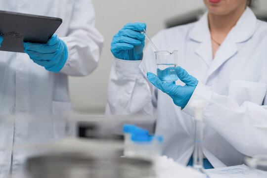 Professional female scientist using glass dropper with blue liquid in beaker for chemical analysis and medical research at modern laboratory, while colleague records data on digital tablet.