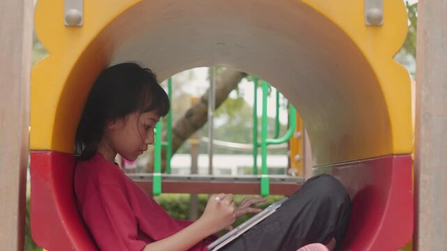 Asian kid drawing digital art on tablet inside a playground tunnel. Selective focus highlights the creative elementary student enjoying casual hobby and relaxing time outdoors with technology.