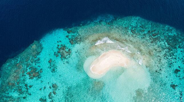 A beautifully detailed photorealistic aerial stock photograph capturing a stunning secluded Caribbean coral reef atoll surrounded by deep blue and vibrant turquoise water.
