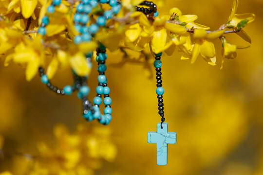 A decorative pendant in the shape of a blue cross on a string of multicolored beads, set against a background of forsythia.