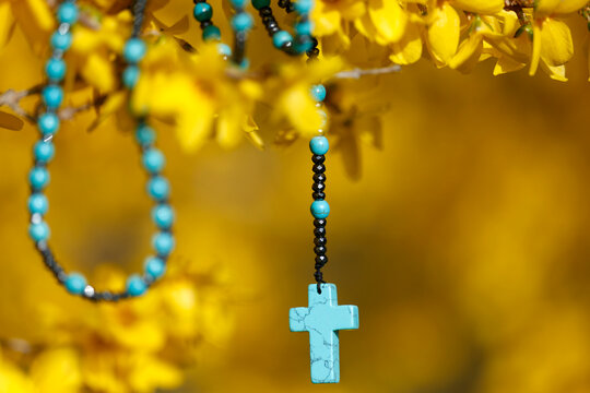 A decorative pendant in the shape of a blue cross on a string of multicolored beads, set against a background of forsythia.