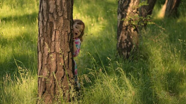 Happy little girl playing hide and seek behind a tree in a sunny park. Cute child laughing, having fun outdoors during spring. Childhood memory, joy, playfulness and exploration concept.