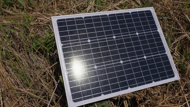 A small solar panel rests on dry yellow straw and grass under bright sunlight in a rural field. Concept of renewable energy, off-grid power, and solar system installation.