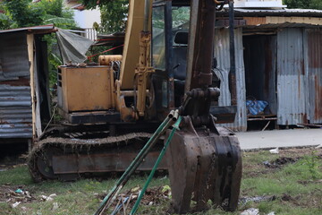 Fototapeta premium A heavy-duty yellow excavator parked on a grassy patch next to rustic corrugated metal shelters and debris.