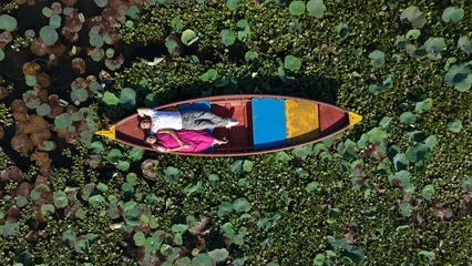 Fotobehang Lotusbloem Aerial drone view of a couple enjoying a colorful boat ride across a scenic lake covered with lotus leaves and aquatic plants, surrounded by lush greenery.  © Vishal