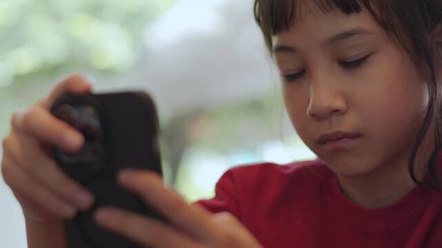 Close up Asian kid seriously focusing on a smartphone at a restaurant. Thai girl intently playing mobile games or watching content. Shot with selective focus and copy space. Digital lifestyle concept.