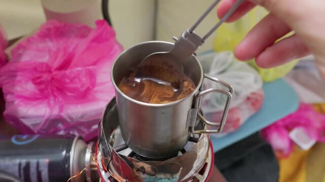 Coffee grounds are scooped into hot water inside a metal camping mug on a portable stove. Concept of outdoor beverage preparation, morning caffeine fix, and camping lifestyle.