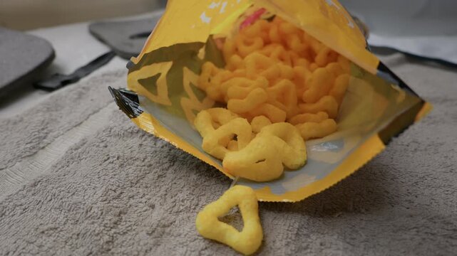 Golden triangle cheese puff snacks spill out from an open yellow bag onto a gray towel surface. Concept of casual snacking, convenience eating, junk food cravings, and quick savory treats.
