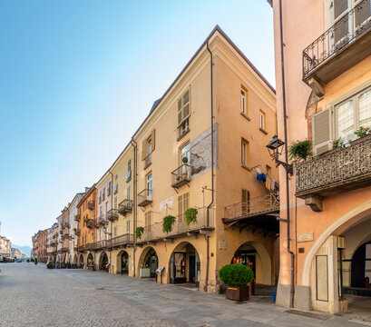 historic Via Roma in Cuneo, Piedmont, Italy, with iconic medieval and neoclassical arcades, traditional pastel-colored residential buildings, stone pavement, and a clear blue sky.