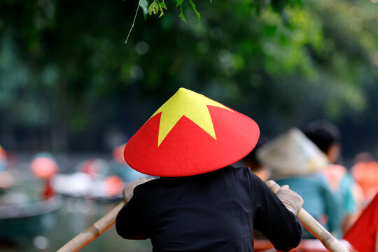 A woman wearing a traditional conical hat adorned with a yellow star on a red background rows a boat on a river in the Ninh Binh region. Trang An, Vietnam.