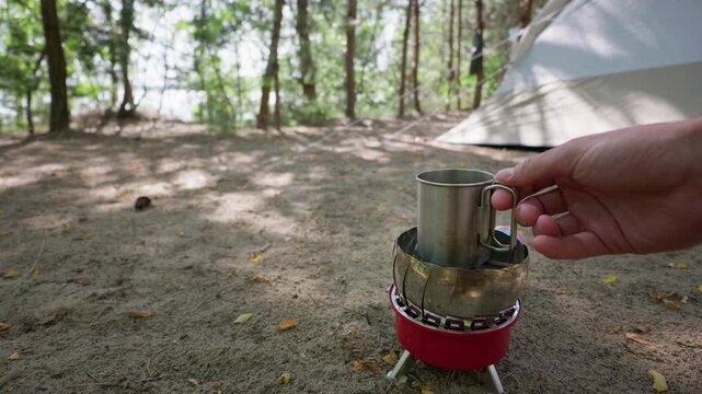 A person holds a stainless steel coffee mug over a small portable burner on sandy ground. A tourist tent rests under tall pine trees during an off-grid outdoor escape in nature.