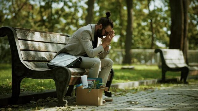 Fired Businessman Sitting On Park Bench With Cardboard Box Personal Belongings. Job Loss And Failure 