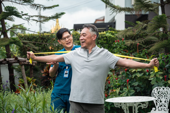 Senior Physical Therapy for an Asian Elderly Man Exercising with a Resistance Band while a Male Caregiver in Blue Medical Scrubs Guides His Movements in a Green Garden