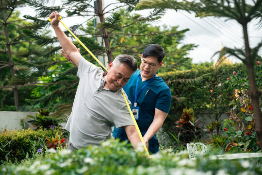 Senior Physical Therapy for an Asian Elderly Man Exercising with a Resistance Band while a Male Caregiver in Blue Medical Scrubs Guides His Movements in a Green Garden