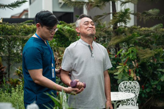 Senior Physical Therapy for an Asian Elderly Man Performing Dumbbell Arm Exercises while a Male Caregiver in Blue Medical Scrubs Provides Support in an Outdoor Garden