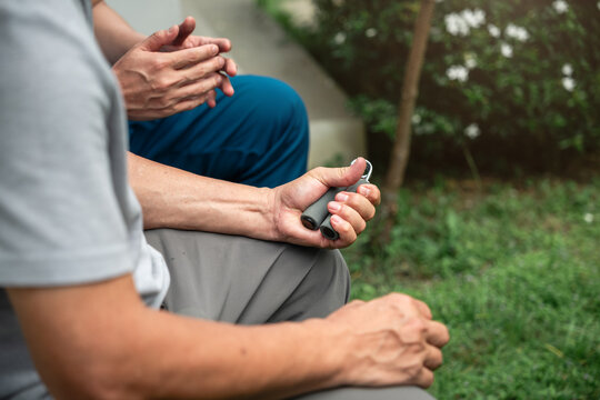 Senior Rehabilitation Close-up of an Asian Elderly Man Using a Hand Grip Exerciser while a Male Caregiver in Blue Medical Scrubs Sits Nearby in an Outdoor Garden