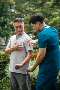 Senior Physical Therapy for an Asian Elderly Man Performing Dumbbell Arm Exercises while a Male Caregiver in Blue Medical Scrubs Provides Support in an Outdoor Garden