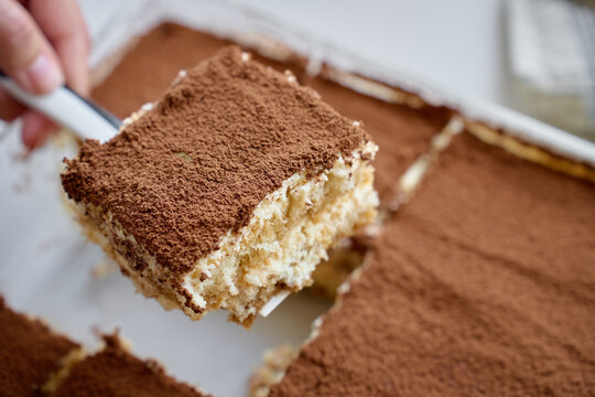 Tiramisu slice is lifted with spatula from dish, showing cocoa topping and cream