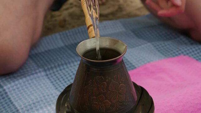 An ornate copper Turkish coffee pot with floral engravings sits atop a compact portable gas stove. The traditional hot beverage brews gently outdoors on a blue patterned picnic blanket.
