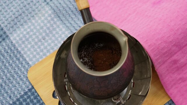 Ground coffee powder sits inside a traditional copper cezve placed on a burner outdoors. Concept of outdoor brewing, camping lifestyle, artisan beverage preparation, and morning routine.