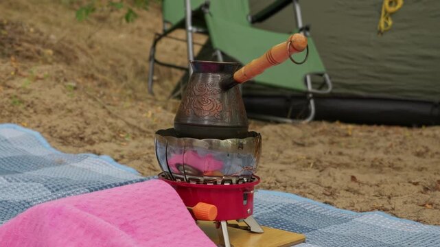 A traditional copper turkish coffee pot sits on a portable gas stove outside a green camping tent. Concept of outdoor living, adventure lifestyle, morning brew, and relaxation in nature.