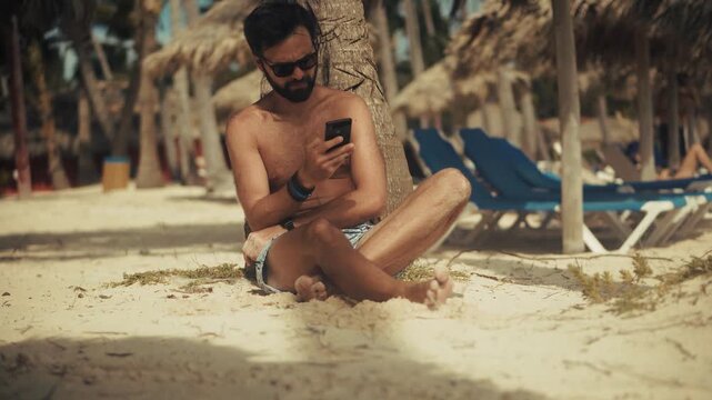 Tourist Using Smartphone Sitting On Sand Under Palm Tree. Tropical Beach Resort. Summer Vacation Leisure 