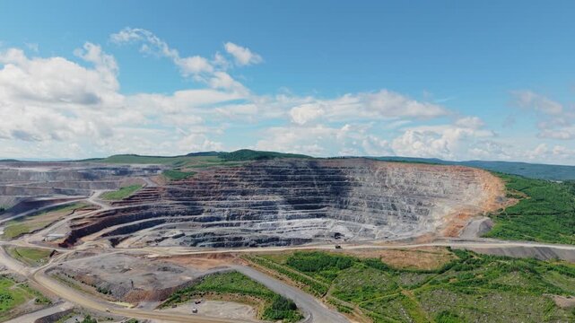 Aerial tracking shot of open pit mine revealing terraced excavation, haul roads and heavy machinery across rural hillside industry landscape.