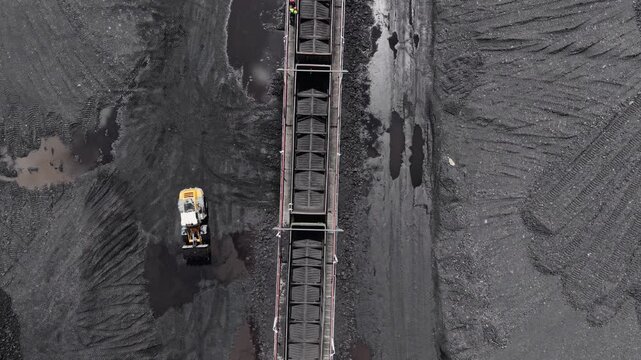 Aerial tracking shot excavator loading coal into railcars at industrial mining yard.