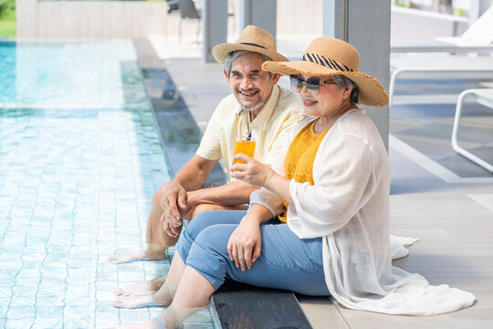 happy asian elderly couple enjoying tropical holiday, dipping feet in swimming pool and drinking refreshment together.
