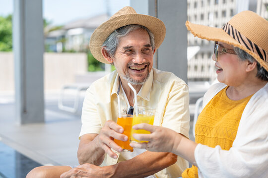 happy senior couple sitting poolside and clinking glass of orange juice together while relaxing on summer vacation at a luxury resort