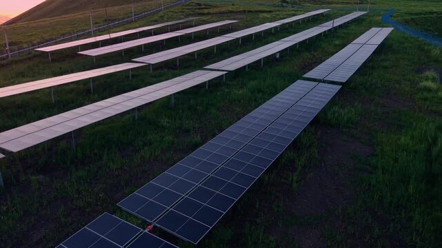 Aerial view panels stretching in rows across grassy hillside solar farm at sunset with slow forward dolly movement and reflective glint, mining industry factory.