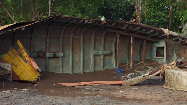 A capsized fishing boat rests on a muddy riverbank after severe flooding, surrounded by debris and damaged infrastructure.