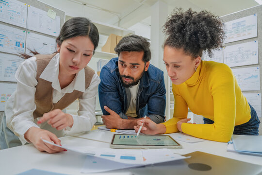 Three colleagues lean over a desk covered in printed charts and a tablet, closely reviewing data together during a focused analysis session in a modern office lined with project boards.